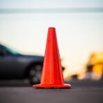 A close-up of a bright orange traffic cone on a city street, with blurred cars in the background.