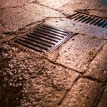 Nighttime shot of wet asphalt road with drainage grates reflecting light.